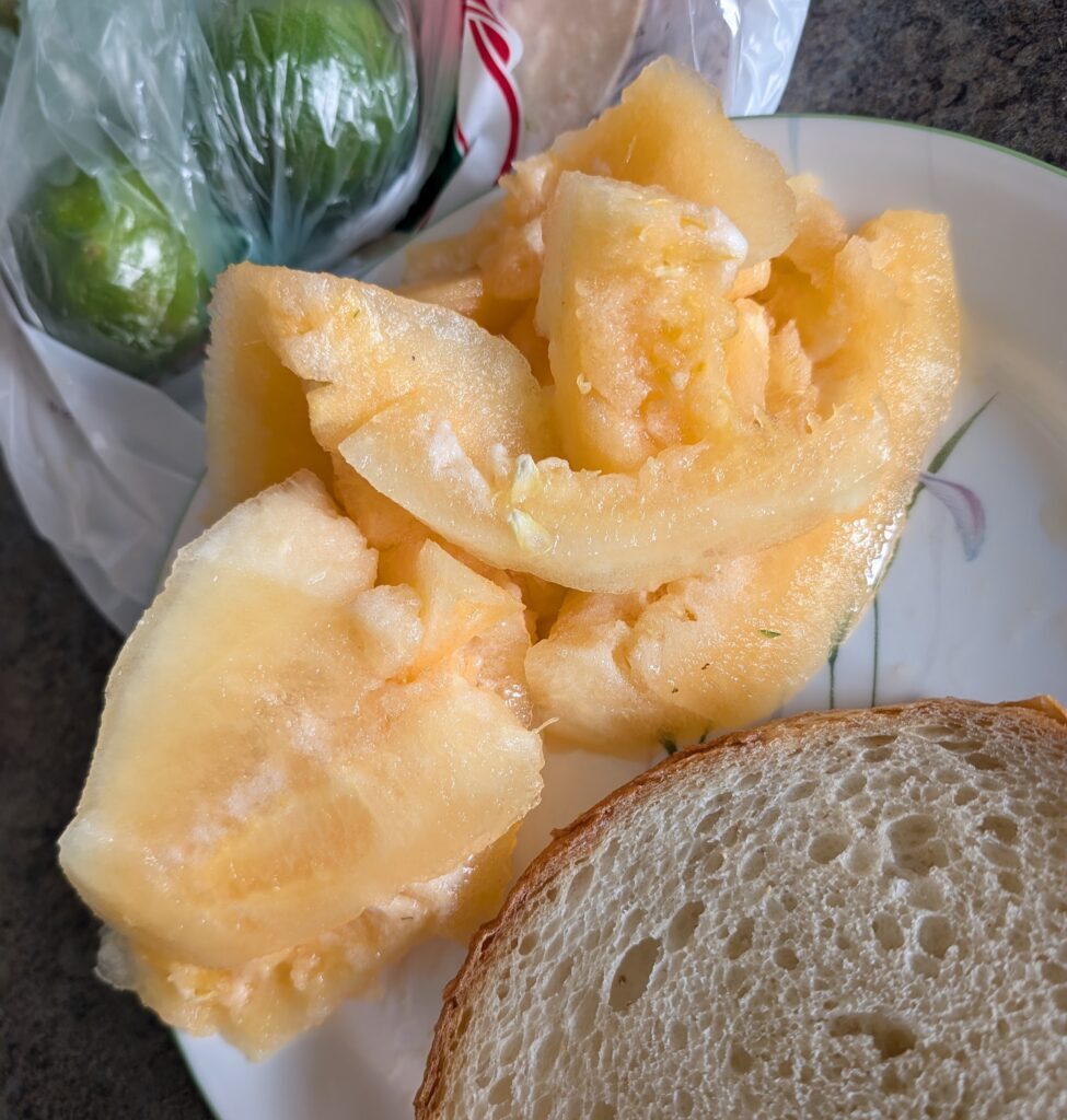 Yellow watermelon flesh carved from the melon, seeds removed, on a plate next to some whole grain bread, which sits on a counter with some limes in a plastic bag nearby