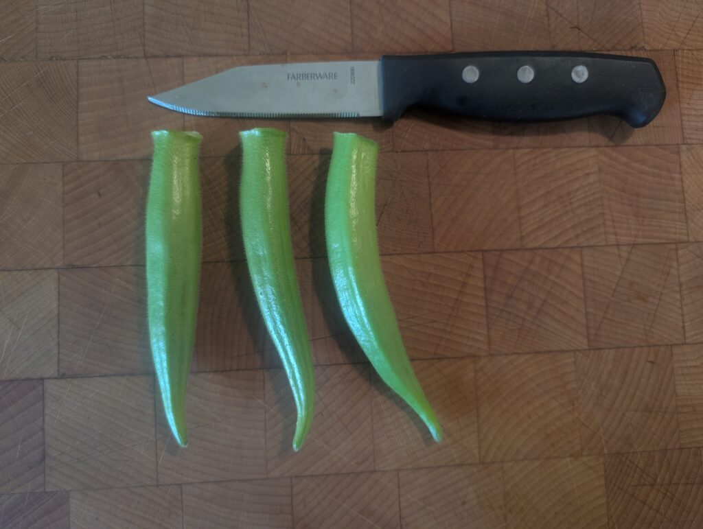 Stewart Zeebest okra pods. A small kitchen knife lying flat horizontal left to right with the blade left on a wooden cutting block. Three okra pods are beneath the knife blade perpendicular to it. They are velvety and green, smooth with no ribs or spines, and slender.